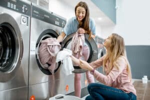 Two Cheerful Girlfriends Having Fun Loading Clothes Into The Washing